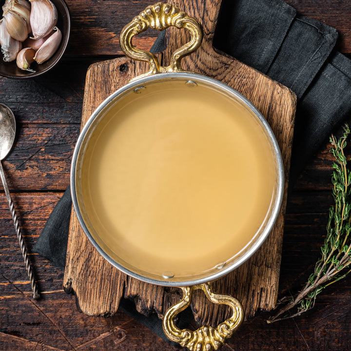 Metal bowl with bone broth on a wooden surface with a spoon and herbs.